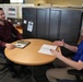 Two men are seated at a round wooden table in an open office environment, engaged in what appears to be a professional meeting or interview. The man on the right, wearing a blue Adidas polo shirt, is gesturing with his hand while holding a pen, suggesting he is actively speaking. The man on the left, in a dark red button-up shirt, is listening attentively with a slight smile.