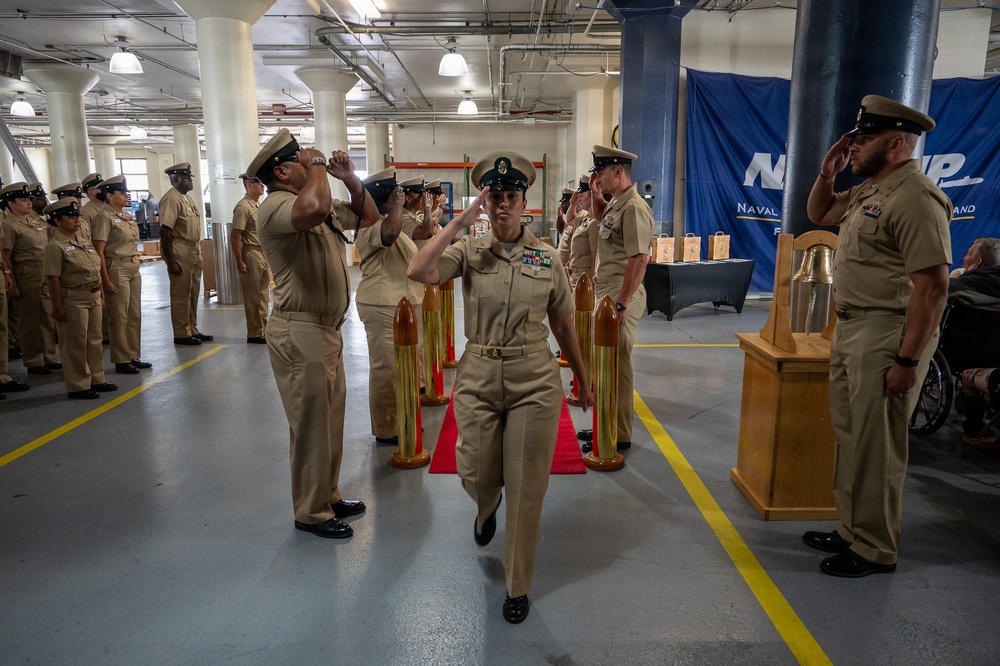 A U.S. Navy chief petty officer pinning or advancement ceremony is taking place in what appears to be a large indoor facility, likely a naval installation. A female sailor in khaki uniform walks through a sideboy honors formation while fellow sailors render salutes on both sides. The setting includes ceremonial elements such as large brass shell casings, a ship's bell, and a blue Naval banner in the background.