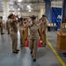 A U.S. Navy chief petty officer pinning or advancement ceremony is taking place in what appears to be a large indoor facility, likely a naval installation. A female sailor in khaki uniform walks through a sideboy honors formation while fellow sailors render salutes on both sides. The setting includes ceremonial elements such as large brass shell casings, a ship's bell, and a blue Naval banner in the background.