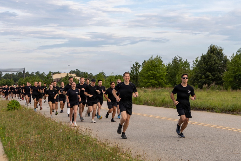 A large group of people dressed uniformly in black t-shirts and black shorts are running together along a two-lane road in what appears to be a organized group run or physical training event. Two individuals lead the formation at the front, with dozens of participants stretching back along the road. The setting appears to be a semi-rural area with trees, grass, and utility poles visible alongside the road.