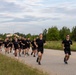 A large group of people dressed uniformly in black t-shirts and black shorts are running together along a two-lane road in what appears to be a organized group run or physical training event. Two individuals lead the formation at the front, with dozens of participants stretching back along the road. The setting appears to be a semi-rural area with trees, grass, and utility poles visible alongside the road.