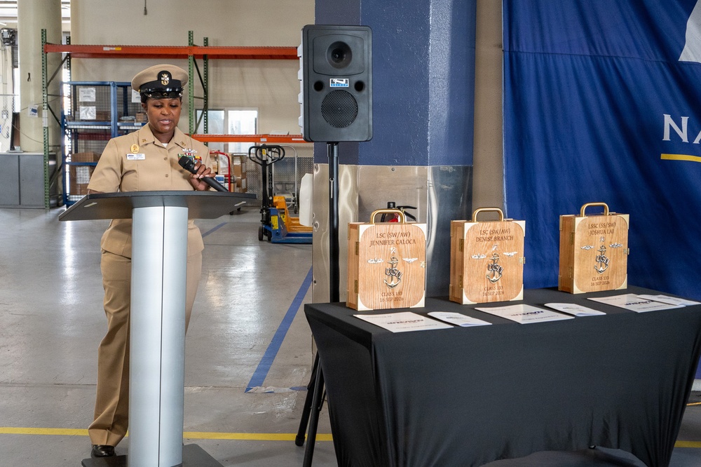 A U.S. Navy chief petty officer in khaki service uniform is speaking at a podium during what appears to be a military advancement or recognition ceremony. Three engraved wooden gift boxes are displayed on a black-draped table, each bearing the names of honorees along with Navy insignia. The event is taking place inside what appears to be a warehouse or industrial facility decorated with a Navy banner.