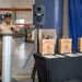 A U.S. Navy chief petty officer in khaki service uniform is speaking at a podium during what appears to be a military advancement or recognition ceremony. Three engraved wooden gift boxes are displayed on a black-draped table, each bearing the names of honorees along with Navy insignia. The event is taking place inside what appears to be a warehouse or industrial facility decorated with a Navy banner.