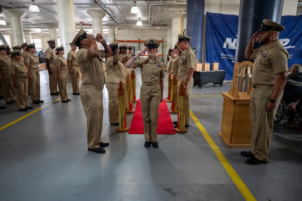 A formal U.S. Navy military ceremony is taking place inside what appears to be a large indoor facility or hangar. Personnel in tan/khaki Navy working uniforms are rendering salutes along a red carpet flanked by brass shell casing stanchions. A blue banner in the background references 'Naval Supply' (NAVSUP), suggesting this is likely a change of command or retirement ceremony.