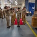 A formal U.S. Navy military ceremony is taking place inside what appears to be a large indoor facility or hangar. Personnel in tan/khaki Navy working uniforms are rendering salutes along a red carpet flanked by brass shell casing stanchions. A blue banner in the background references 'Naval Supply' (NAVSUP), suggesting this is likely a change of command or retirement ceremony.