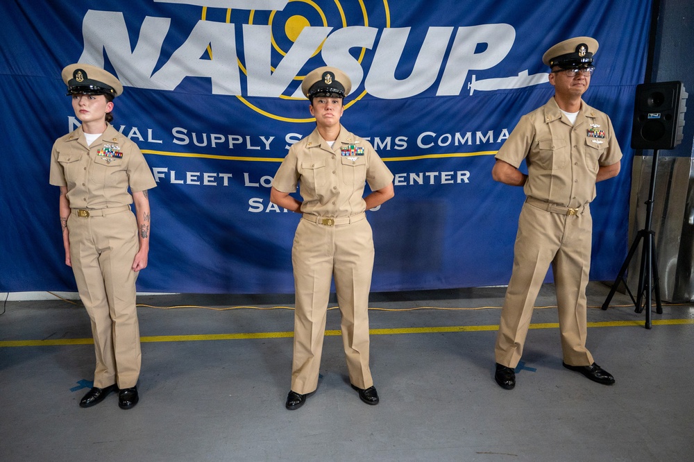 Three U.S. Navy personnel in khaki service uniforms stand at attention in front of a NAVSUP (Naval Supply Systems Command) Fleet Logistics Center banner. The individuals display various service ribbons and insignia on their uniforms, suggesting different ranks and service histories. The setting appears to be an official military ceremony or formal event held in a warehouse or hangar-type facility.