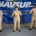 Three U.S. Navy personnel in khaki service uniforms stand at attention in front of a NAVSUP (Naval Supply Systems Command) Fleet Logistics Center banner. The individuals display various service ribbons and insignia on their uniforms, suggesting different ranks and service histories. The setting appears to be an official military ceremony or formal event held in a warehouse or hangar-type facility.