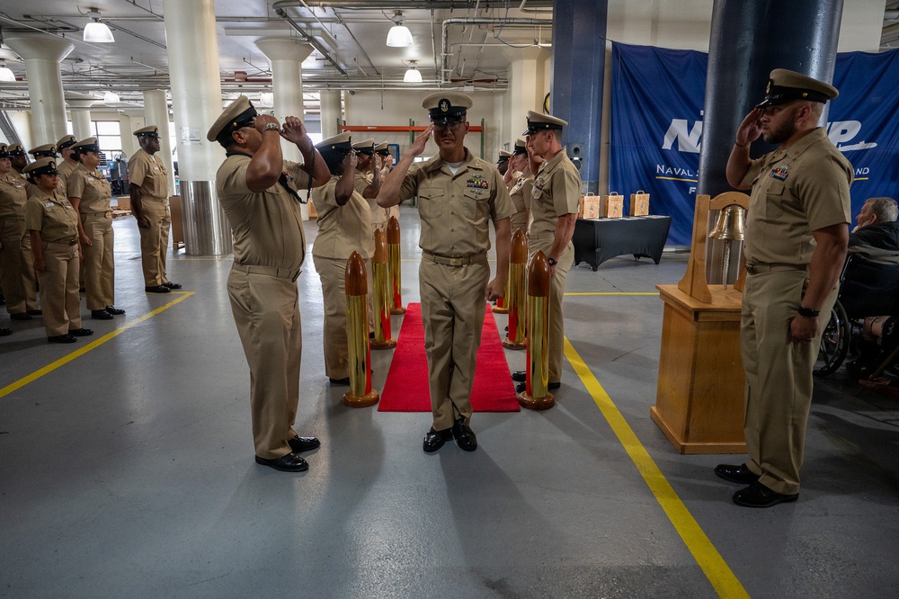A formal U.S. Navy ceremony is taking place inside what appears to be a large indoor facility, likely a hangar or warehouse. Personnel in khaki service uniforms are rendering salutes along a red carpet flanked by decorative brass shell casings, suggesting a sideboy or honors ceremony. A blue banner in the background reads 'Naval Support' indicating this is a Naval Support Activity installation.