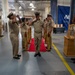 A formal U.S. Navy ceremony is taking place inside what appears to be a large indoor facility, likely a hangar or warehouse. Personnel in khaki service uniforms are rendering salutes along a red carpet flanked by decorative brass shell casings, suggesting a sideboy or honors ceremony. A blue banner in the background reads 'Naval Support' indicating this is a Naval Support Activity installation.