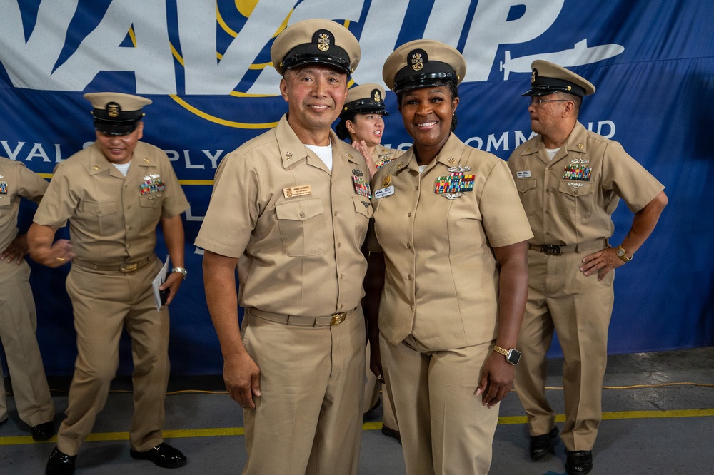 A group of U.S. Navy personnel in tan/khaki service uniforms are gathered in front of a blue banner reading 'Naval Supply Systems Command.' Two chief petty officers in the foreground are posing together and smiling for the camera. The setting appears to be an official military ceremony or event.