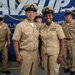 A group of U.S. Navy personnel in tan/khaki service uniforms are gathered in front of a blue banner reading 'Naval Supply Systems Command.' Two chief petty officers in the foreground are posing together and smiling for the camera. The setting appears to be an official military ceremony or event.