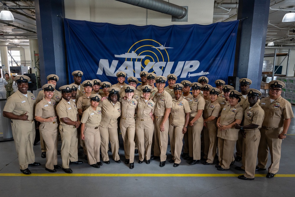 A large group of U.S. Navy personnel in tan/khaki service uniforms pose for a formal group photograph in front of a NAVSUP (Naval Supply Systems Command) banner. The setting appears to be a warehouse or logistics facility, consistent with NAVSUP's supply chain mission. The group includes both male and female sailors of diverse backgrounds, suggesting a unit or command photo.