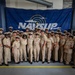 A large group of U.S. Navy personnel in tan/khaki service uniforms pose for a formal group photograph in front of a NAVSUP (Naval Supply Systems Command) banner. The setting appears to be a warehouse or logistics facility, consistent with NAVSUP's supply chain mission. The group includes both male and female sailors of diverse backgrounds, suggesting a unit or command photo.