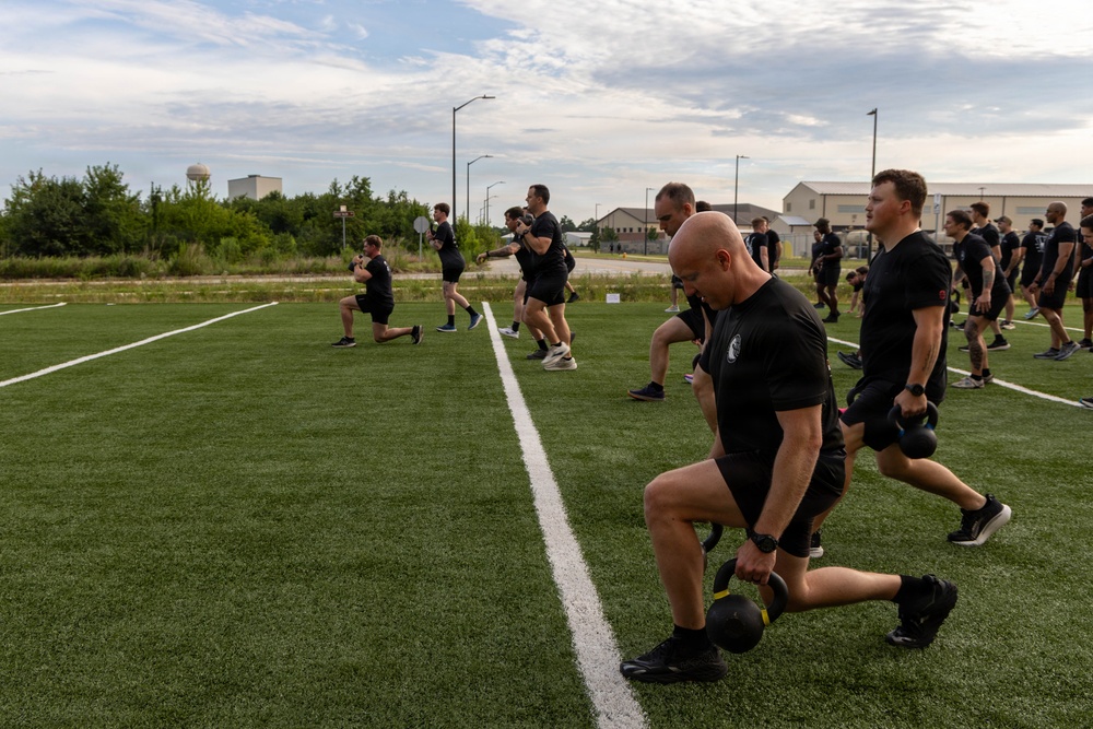 A large group of individuals dressed in matching black athletic attire are performing kettlebell lunges on an artificial turf field. The exercise appears to be a coordinated group physical training session, likely military or law enforcement in nature given the uniformity of dress and disciplined formation. The setting is an outdoor sports field with buildings and a water tower visible in the background.