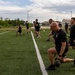 A large group of individuals dressed in matching black athletic attire are performing kettlebell lunges on an artificial turf field. The exercise appears to be a coordinated group physical training session, likely military or law enforcement in nature given the uniformity of dress and disciplined formation. The setting is an outdoor sports field with buildings and a water tower visible in the background.