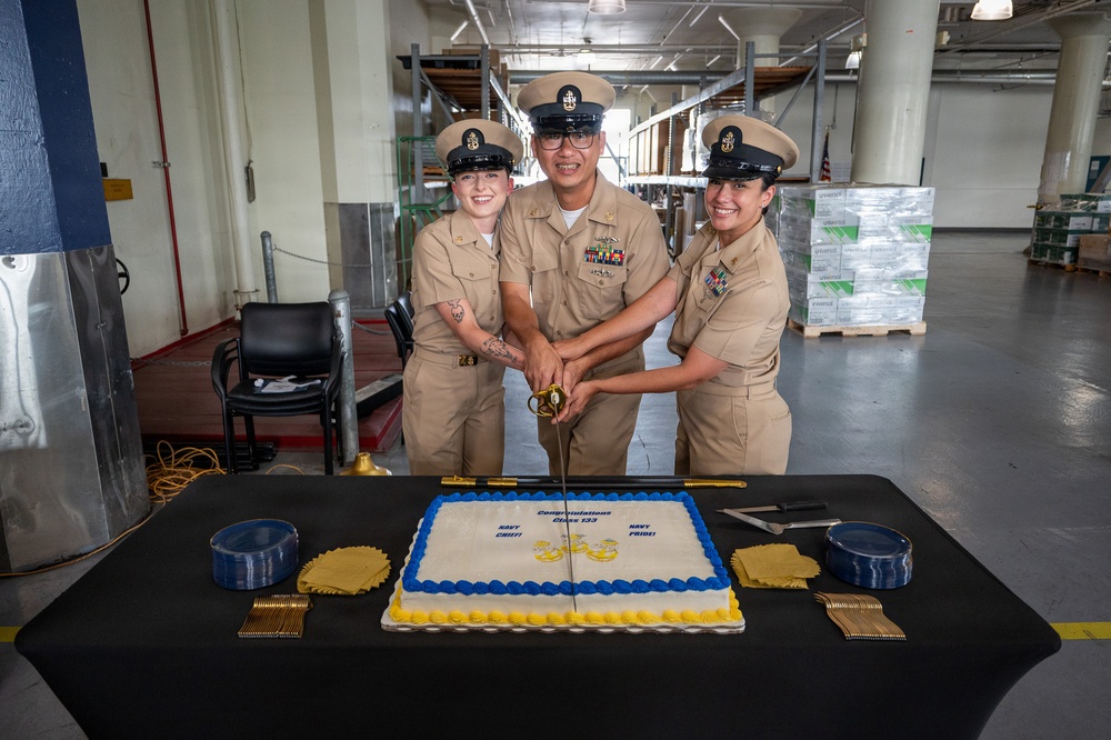Three U.S. Navy Chief Petty Officers in Service Khaki uniforms are cutting a ceremonial cake with a sword at what appears to be a Chief Petty Officer pinning or advancement ceremony. The cake reads 'Congratulations Class 133' with 'Navy Chief' and 'Navy Pride' inscriptions, decorated in Navy blue and gold colors. The event is taking place inside a large warehouse or industrial facility.