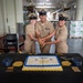 Three U.S. Navy Chief Petty Officers in Service Khaki uniforms are cutting a ceremonial cake with a sword at what appears to be a Chief Petty Officer pinning or advancement ceremony. The cake reads 'Congratulations Class 133' with 'Navy Chief' and 'Navy Pride' inscriptions, decorated in Navy blue and gold colors. The event is taking place inside a large warehouse or industrial facility.