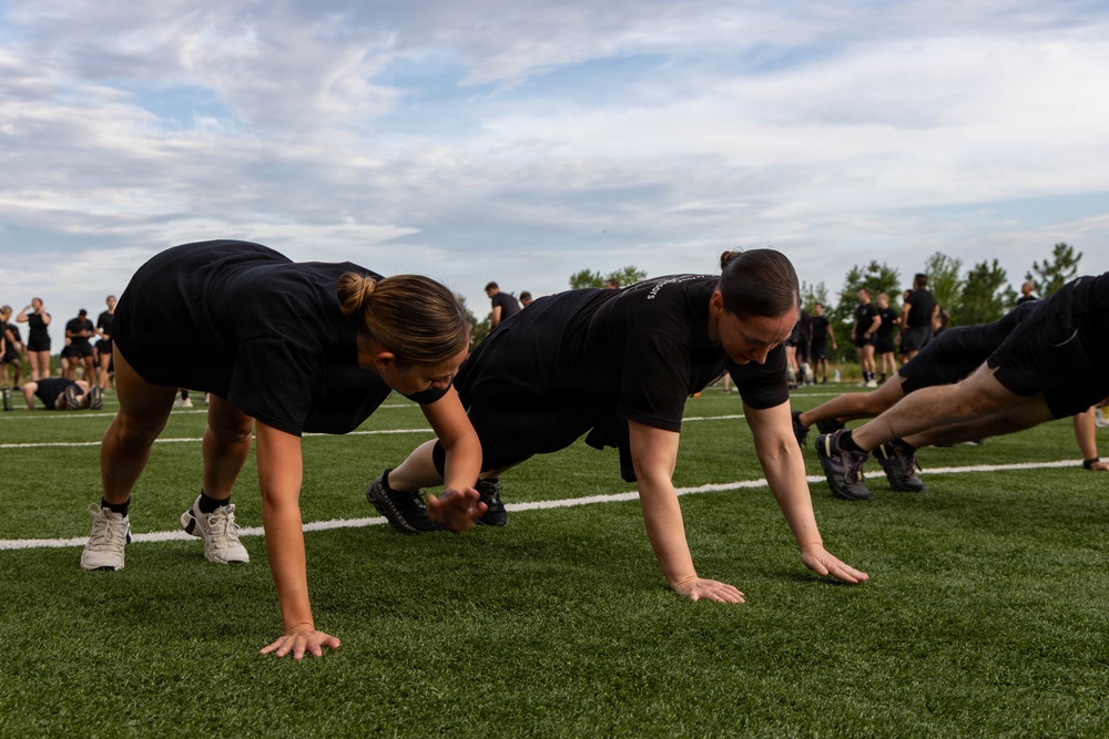 A group of individuals dressed in matching black t-shirts and shorts are performing push-ups on a green artificial turf field. The scene appears to be a structured physical training session, likely military, law enforcement, or athletic in nature, with multiple participants visible in the foreground and background. The setting is outdoors under a partly cloudy sky with trees visible in the distance.