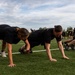 A group of individuals dressed in matching black t-shirts and shorts are performing push-ups on a green artificial turf field. The scene appears to be a structured physical training session, likely military, law enforcement, or athletic in nature, with multiple participants visible in the foreground and background. The setting is outdoors under a partly cloudy sky with trees visible in the distance.