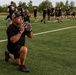 A large group of individuals dressed uniformly in black athletic wear are participating in an outdoor fitness training session on a synthetic turf field. The foreground shows a middle-aged bearded man in a trucker cap performing a lunge while holding a medicine ball at chest level. In the background, participants are engaged in various exercises including kettlebell work and running drills.
