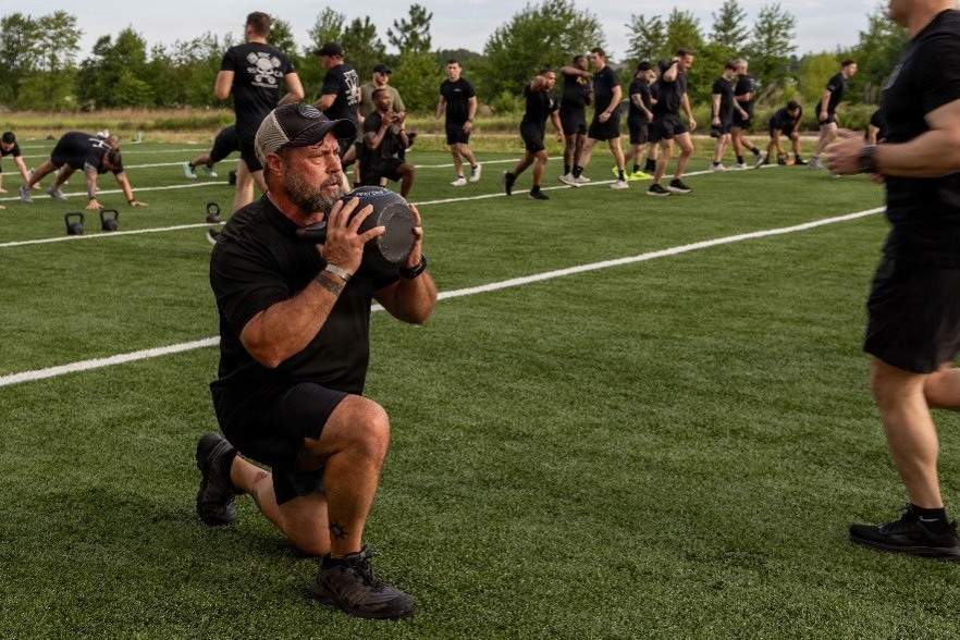 A large group of individuals dressed uniformly in black athletic wear are participating in an outdoor fitness training session on a synthetic turf field. The foreground shows a middle-aged bearded man in a trucker cap performing a lunge while holding a medicine ball at chest level. In the background, participants are engaged in various exercises including kettlebell work and running drills.