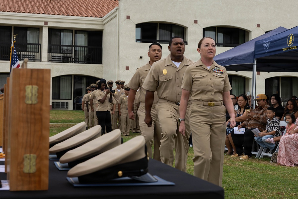 A U.S. Navy military ceremony is taking place outdoors, likely a Chief Petty Officer (CPO) pinning or advancement ceremony. Personnel in khaki service uniforms are marching in formation toward a table displaying cover (hats) and wooden presentation boxes. Civilian family members and guests are seated to the right observing the proceedings.