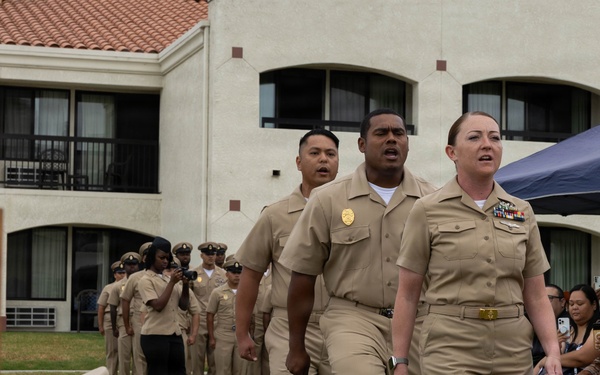 A U.S. Navy military ceremony is taking place outdoors, likely a Chief Petty Officer (CPO) pinning or advancement ceremony. Personnel in khaki service uniforms are marching in formation toward a table displaying cover (hats) and wooden presentation boxes. Civilian family members and guests are seated to the right observing the proceedings.