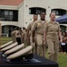 A U.S. Navy military ceremony is taking place outdoors, likely a Chief Petty Officer (CPO) pinning or advancement ceremony. Personnel in khaki service uniforms are marching in formation toward a table displaying cover (hats) and wooden presentation boxes. Civilian family members and guests are seated to the right observing the proceedings.