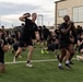 A large group of individuals dressed in black military-style PT (physical training) uniforms are conducting an outdoor group fitness session on a turf field. Participants are performing exercises with kettlebells and weighted objects, including lunges and carries. The setting appears to be a military installation based on the visible 'ARMY' text on clothing and the industrial buildings in the background.