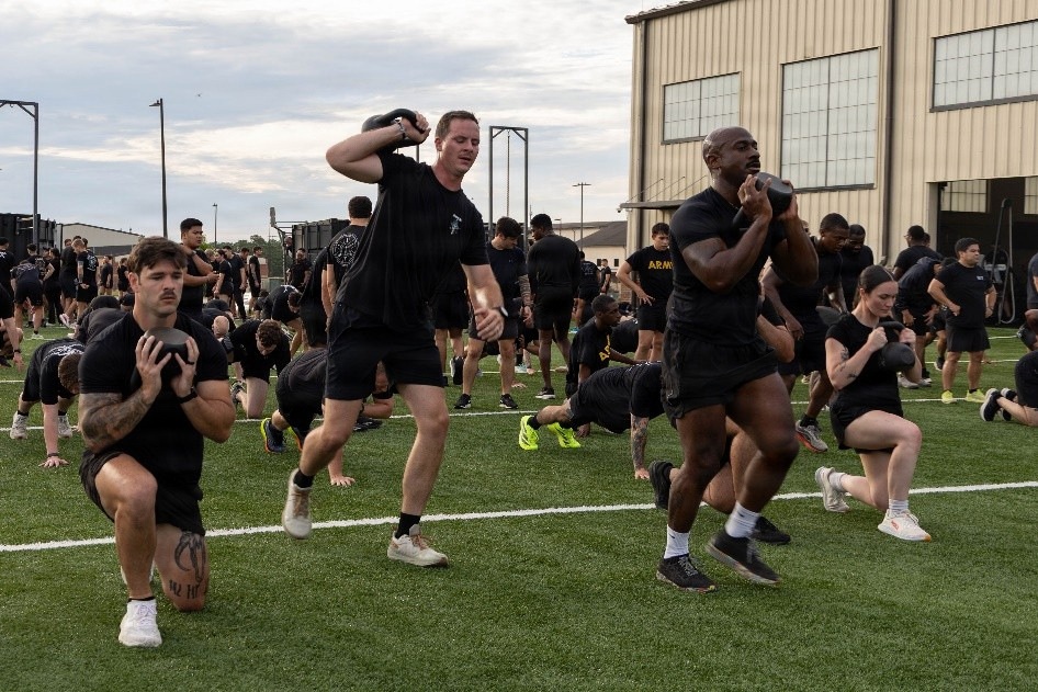A large group of individuals dressed in black military-style PT (physical training) uniforms are conducting an outdoor group fitness session on a turf field. Participants are performing exercises with kettlebells and weighted objects, including lunges and carries. The setting appears to be a military installation based on the visible 'ARMY' text on clothing and the industrial buildings in the background.