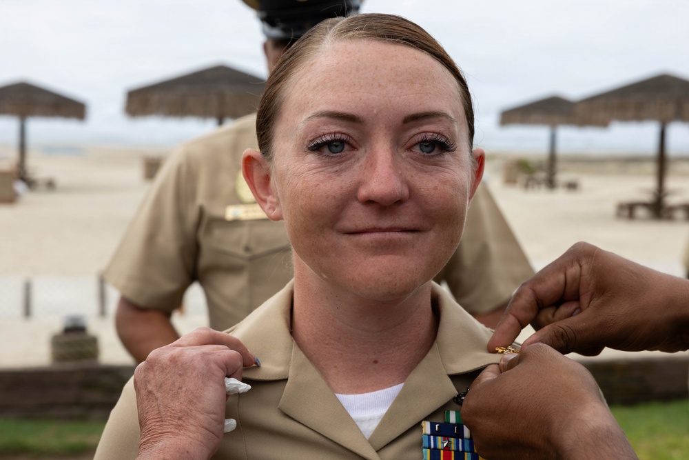 A female U.S. Navy service member is being pinned with insignia or rank devices on her collar during what appears to be a military promotion or award ceremony. She is wearing the standard tan/khaki Navy Service Uniform (NSU) with a ribbon rack visible on her chest. The ceremony appears to be taking place outdoors near a beach area, with thatched-roof shade structures visible in the background.