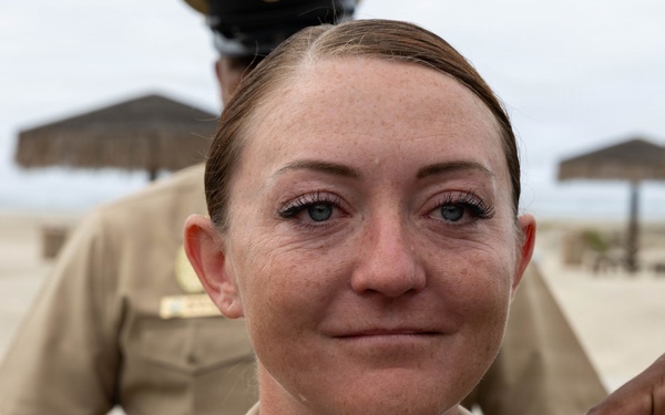 A female U.S. Navy service member is being pinned with insignia or rank devices on her collar during what appears to be a military promotion or award ceremony. She is wearing the standard tan/khaki Navy Service Uniform (NSU) with a ribbon rack visible on her chest. The ceremony appears to be taking place outdoors near a beach area, with thatched-roof shade structures visible in the background.