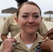 A female U.S. Navy service member is being pinned with insignia or rank devices on her collar during what appears to be a military promotion or award ceremony. She is wearing the standard tan/khaki Navy Service Uniform (NSU) with a ribbon rack visible on her chest. The ceremony appears to be taking place outdoors near a beach area, with thatched-roof shade structures visible in the background.