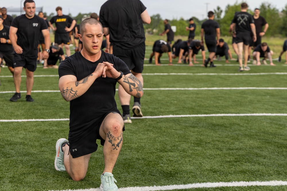 A group of individuals dressed in black athletic uniforms are participating in a physical training session on a turf field. The foreground subject, a tattooed male, is performing a lunge exercise while checking his smartwatch. In the background, multiple participants are engaged in various exercises including push-ups and stretching.