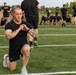 A group of individuals dressed in black athletic uniforms are participating in a physical training session on a turf field. The foreground subject, a tattooed male, is performing a lunge exercise while checking his smartwatch. In the background, multiple participants are engaged in various exercises including push-ups and stretching.