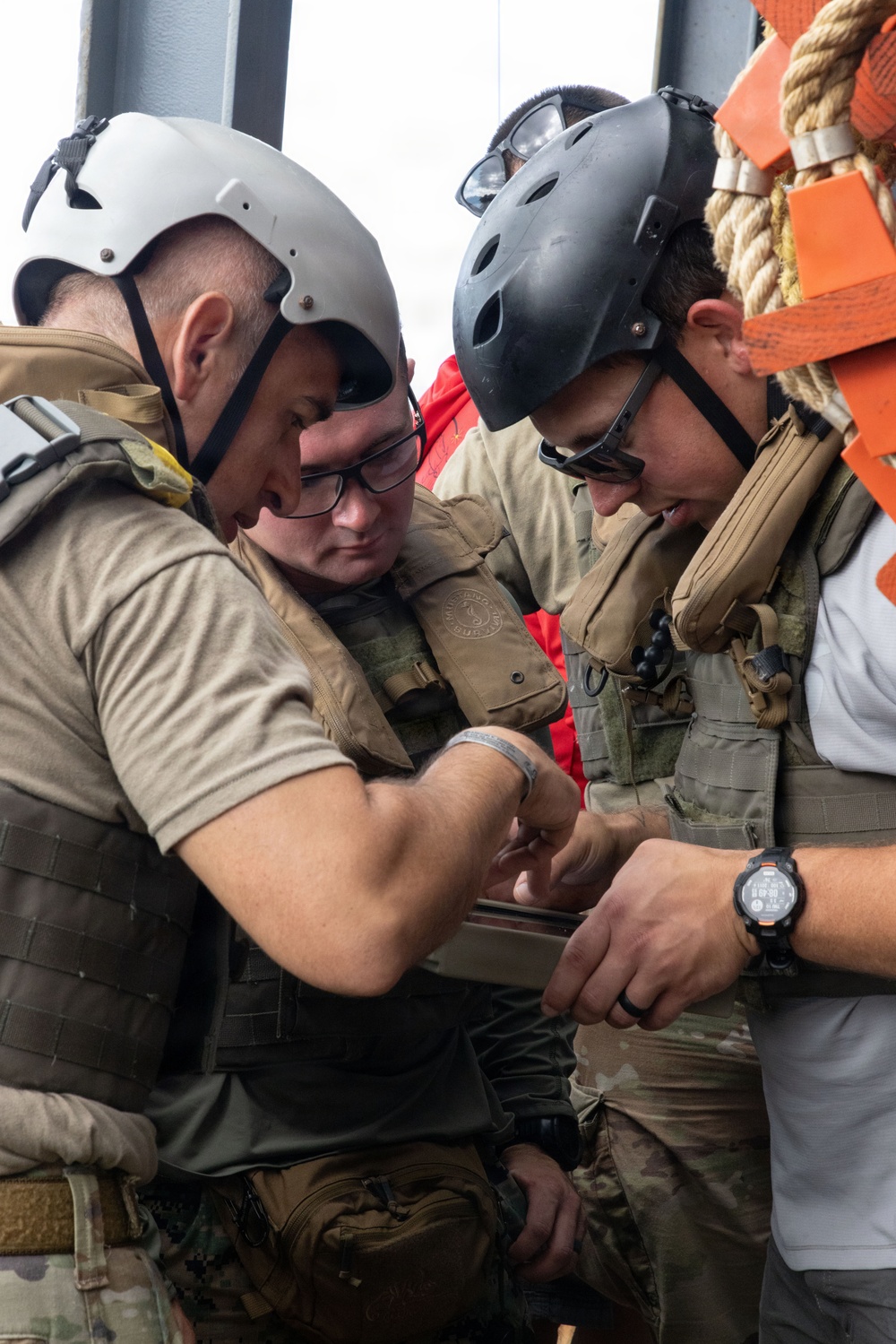 Three individuals in tactical gear and helmets are gathered closely together, appearing to examine or work on a piece of equipment. They are wearing tactical vests, helmets with goggles, and are positioned near what appears to be orange safety equipment and rope on what may be a vessel or platform. The scene suggests a military, coast guard, or maritime security operation.