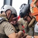 Three individuals in tactical gear and helmets are gathered closely together, appearing to examine or work on a piece of equipment. They are wearing tactical vests, helmets with goggles, and are positioned near what appears to be orange safety equipment and rope on what may be a vessel or platform. The scene suggests a military, coast guard, or maritime security operation.