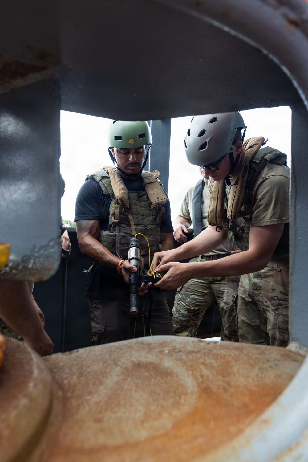 Two military or tactical personnel wearing helmets and plate carriers are working together on a piece of equipment, viewed through a circular metal opening or hatch. One individual holds a tool or device with a yellow cable attached while the other manipulates it. The setting appears to be an industrial or maritime structure with visible rust and metal framework.