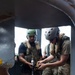 Two military or tactical personnel wearing helmets and plate carriers are working together on a piece of equipment, viewed through a circular metal opening or hatch. One individual holds a tool or device with a yellow cable attached while the other manipulates it. The setting appears to be an industrial or maritime structure with visible rust and metal framework.