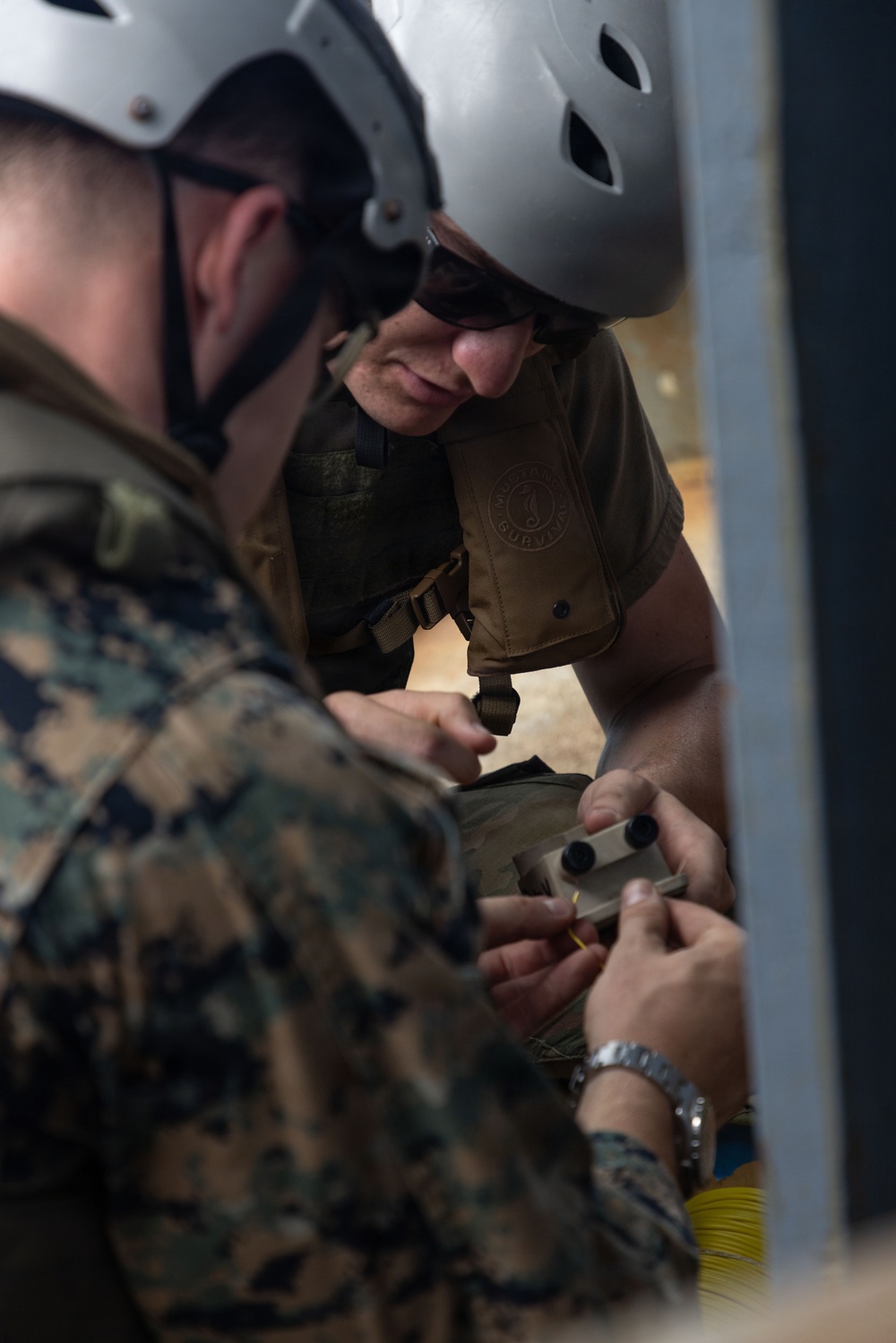 Two individuals in military/tactical gear are closely examining or working with a small device or component. One person wears a white helmet with ventilation holes and sunglasses along with a tan tactical vest, while the other wears a military camouflage uniform. They appear to be collaborating on a technical task involving a small handheld object.
