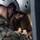 Two individuals in military/tactical gear are closely examining or working with a small device or component. One person wears a white helmet with ventilation holes and sunglasses along with a tan tactical vest, while the other wears a military camouflage uniform. They appear to be collaborating on a technical task involving a small handheld object.