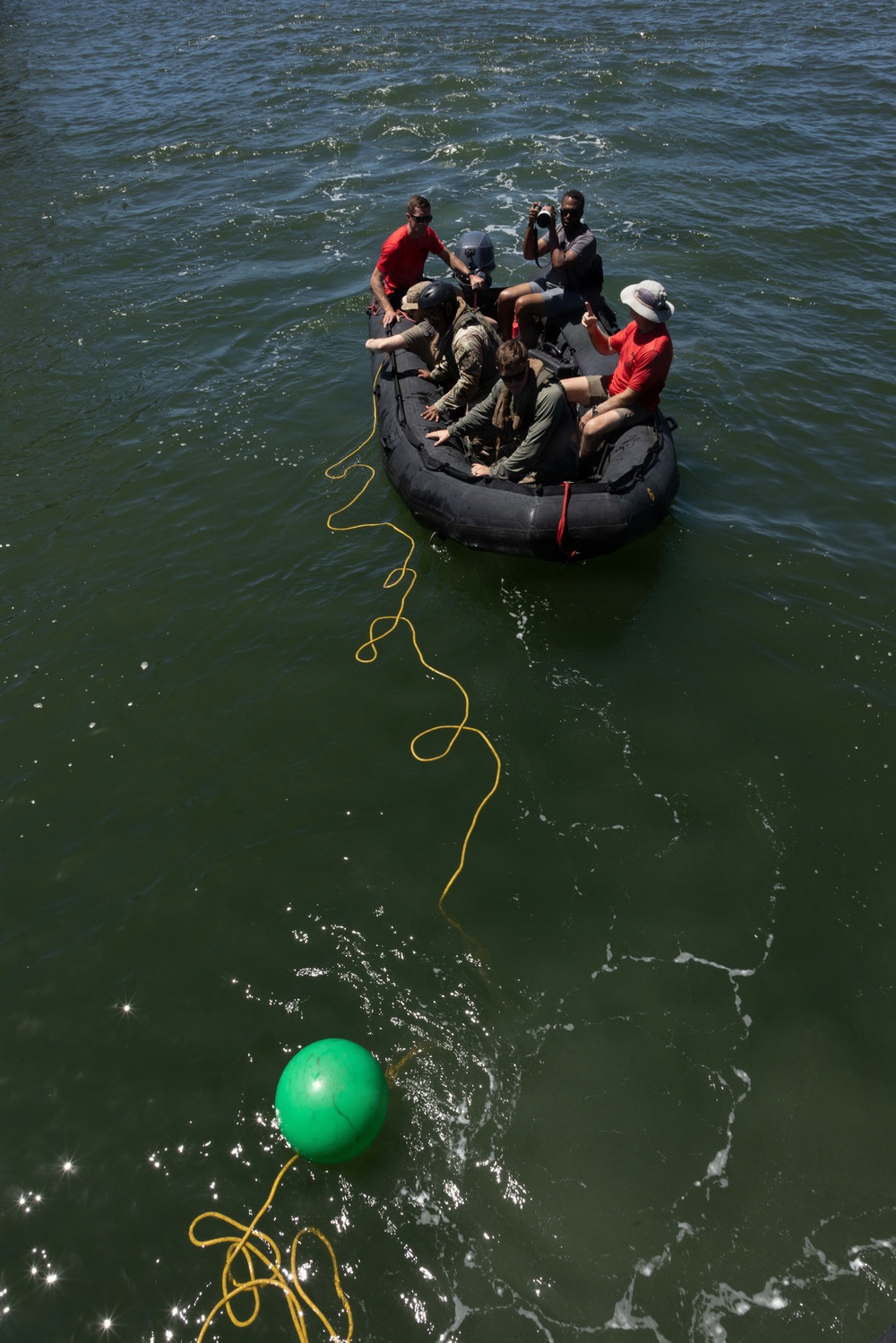 A group of approximately five to six individuals are aboard a black inflatable rubber boat (IRB/Zodiac-style) on open water. A yellow rope extends from the boat toward a green spherical buoy or marker floating in the water below. One person appears to be operating a camera, suggesting this may be a documented training exercise or operation.
