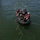 A group of approximately five to six individuals are aboard a black inflatable rubber boat (IRB/Zodiac-style) on open water. A yellow rope extends from the boat toward a green spherical buoy or marker floating in the water below. One person appears to be operating a camera, suggesting this may be a documented training exercise or operation.