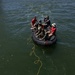 A group of approximately five to six individuals are aboard a black inflatable rubber boat (IRB/Zodiac-style) on open water. A yellow rope extends from the boat toward a green spherical buoy or marker floating in the water below. One person appears to be operating a camera, suggesting this may be a documented training exercise or operation.