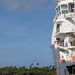 A military or law enforcement boarding operation is underway, with personnel climbing a rope/Jacob's ladder up the side of a large white vessel. A rigid inflatable boat (RIB) with several occupants in red and tactical gear is positioned alongside the ship. The scene appears to take place in a tropical coastal waterway with palm trees visible in the background.