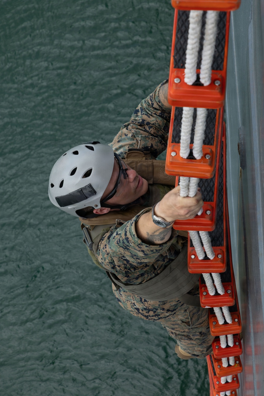 A military personnel member in MARPAT digital camouflage uniform is climbing a Jacob's ladder (rope-and-rung boarding ladder) mounted on the side of a vessel above open water. The individual is wearing a white safety helmet and protective eyewear, gripping the orange-framed ladder with white rope rungs. This appears to be a maritime boarding or ship-climbing training exercise.