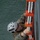 A military personnel member in MARPAT digital camouflage uniform is climbing a Jacob's ladder (rope-and-rung boarding ladder) mounted on the side of a vessel above open water. The individual is wearing a white safety helmet and protective eyewear, gripping the orange-framed ladder with white rope rungs. This appears to be a maritime boarding or ship-climbing training exercise.