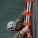 A military personnel member in MARPAT digital camouflage uniform is climbing a Jacob's ladder (rope-and-rung boarding ladder) mounted on the side of a vessel above open water. The individual is wearing a white safety helmet and protective eyewear, gripping the orange-framed ladder with white rope rungs. This appears to be a maritime boarding or ship-climbing training exercise.