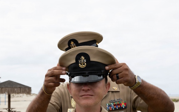 A U.S. Navy Chief Petty Officer is having a combination cover (cap) placed on their head by another person standing behind them, in what appears to be a promotion or pinning ceremony. Both individuals are wearing U.S. Navy Service Uniform Bravo (tan/khaki). The setting appears to be an outdoor beach or coastal military installation.