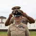 A U.S. Navy Chief Petty Officer is having a combination cover (cap) placed on their head by another person standing behind them, in what appears to be a promotion or pinning ceremony. Both individuals are wearing U.S. Navy Service Uniform Bravo (tan/khaki). The setting appears to be an outdoor beach or coastal military installation.